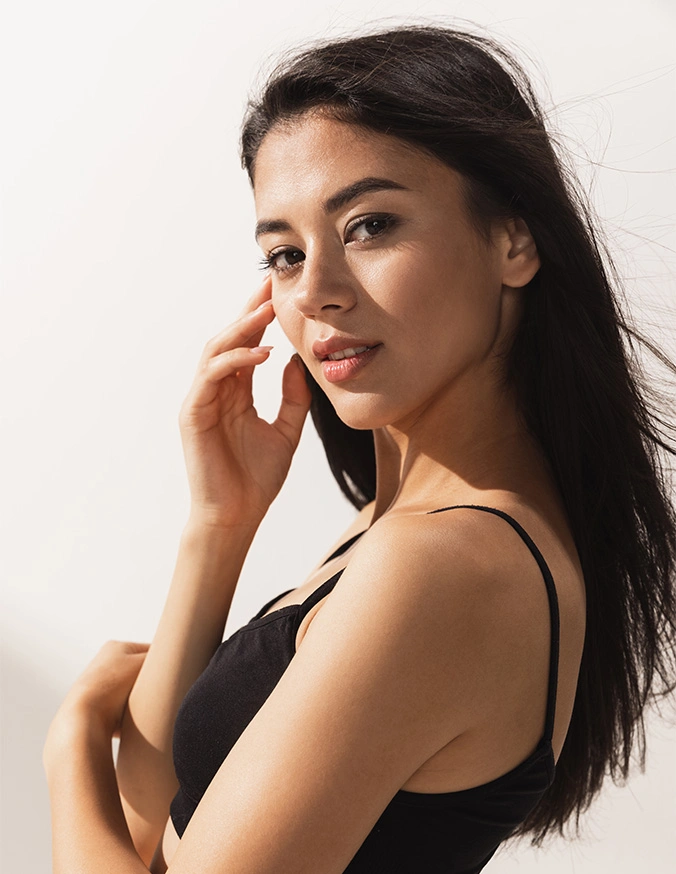 Beautiful woman with long dark hair touching face, wearing black tank top against light background - Buccal fat removal in San Antonio, TX