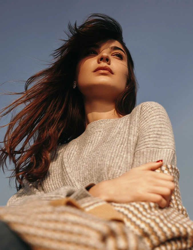 Woman in beige knit sweater with windswept hair against blue sky, arms crossed in confident pose - Lip fillers in San Antonio, TX