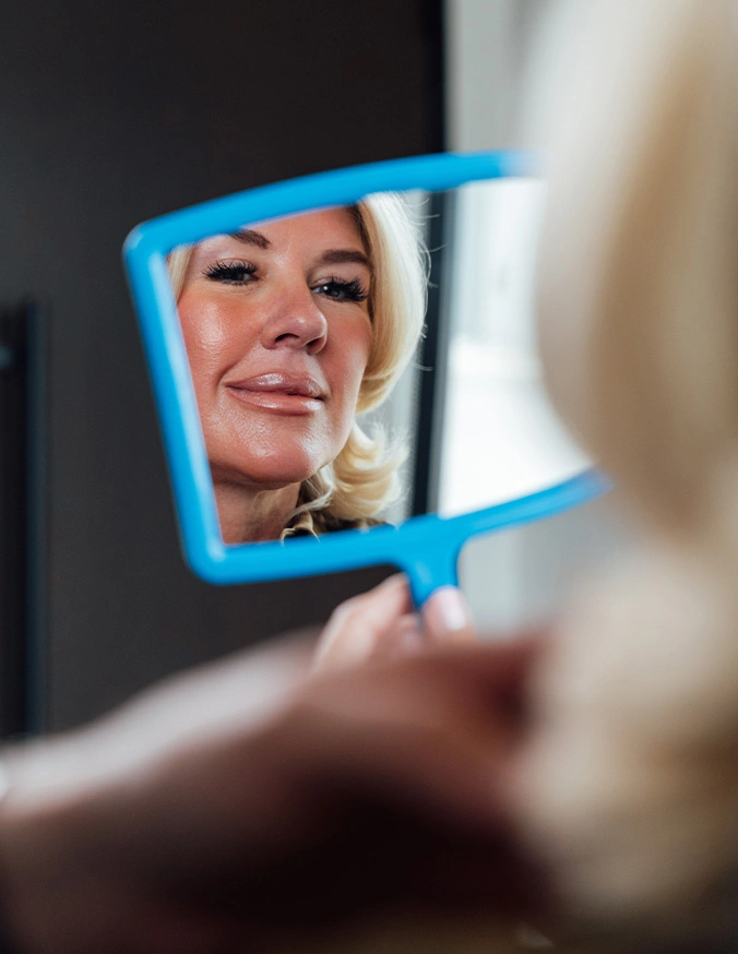 Smiling woman looking at her reflection in a handheld blue mirror while checking her appearance - Neck liposuction in San Antonio, TX