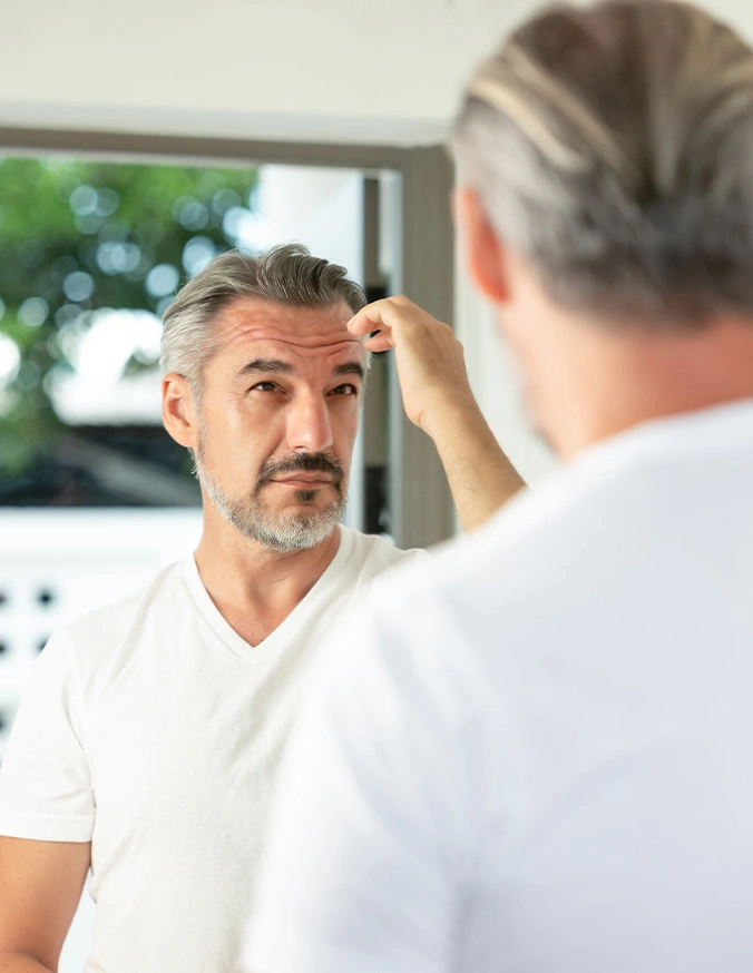 Man examining his gray hair in bathroom mirror while checking hairline and scalp - Neograft in San Antonio, TX