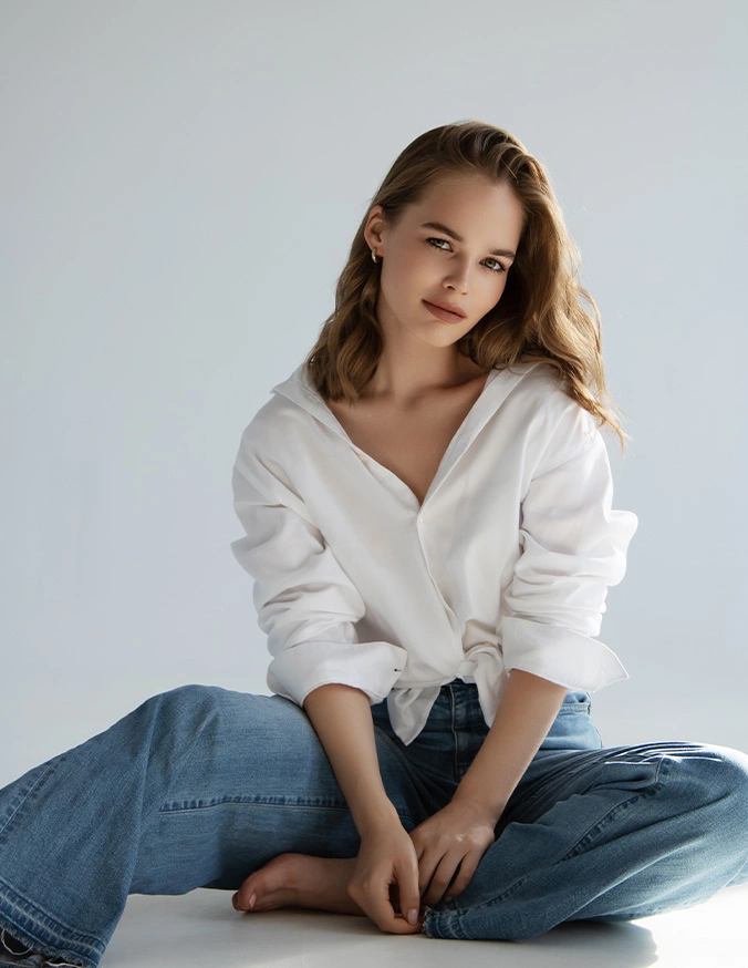 Young woman in white sweater and jeans sitting cross-legged, smiling at camera against neutral background - Otoplasty in San Antonio, TX
