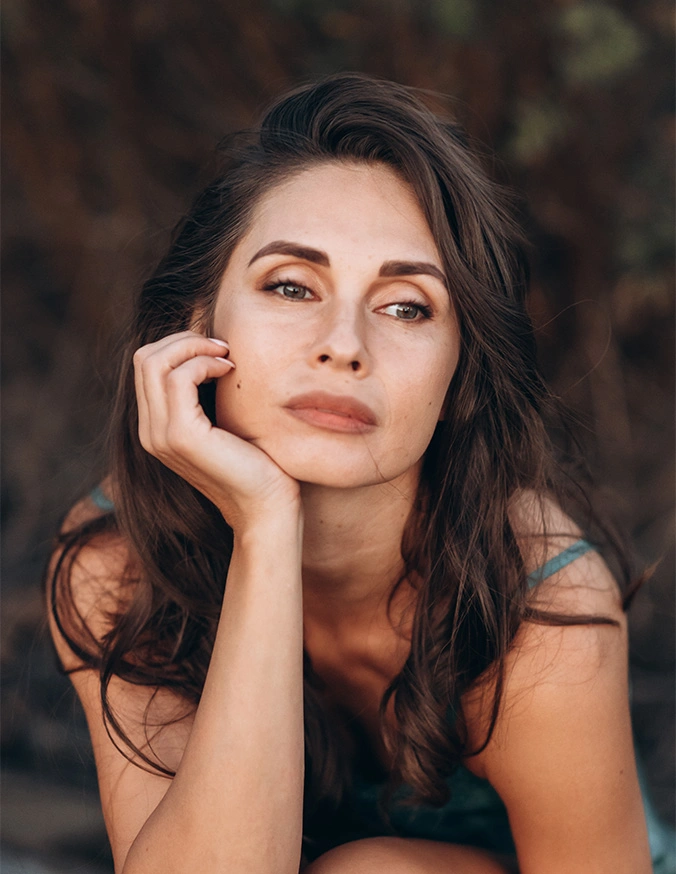 Beautiful young woman with long dark hair resting chin on hand in natural outdoor portrait - Sculptra in San Antonio, TX