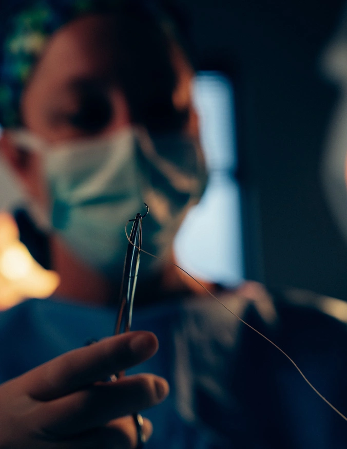 Dr Rokosz threading needle in dim lighting with blurred face in background, close-up of hands doing needlework - Trauma reconstruction in San Antonio, TX