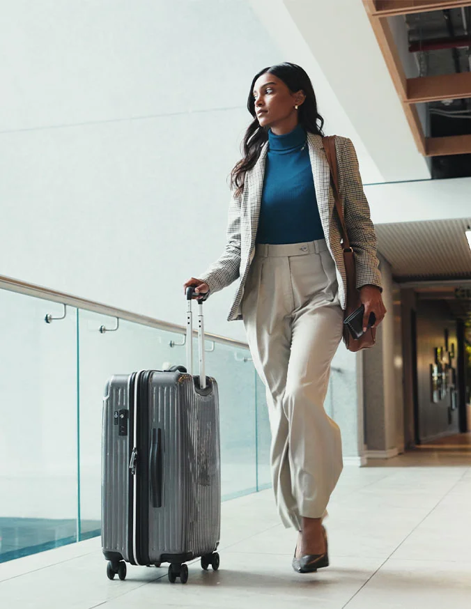 Beautiful Indian woman in a blue turtle neck and white pants and blazer, rolling a silver suitcase through an airport terminal, glancing to the side - Out of town patient resources in San Antonio, TX